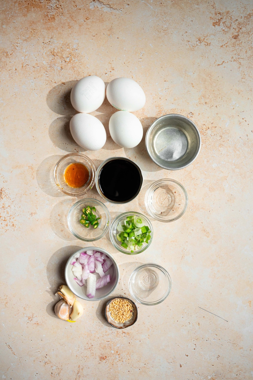 Four eggs, a bowl of soy sauce, bowl, chili, oil, bowl, scallions, bowl, chopped onion, a bowl of garlic, and a bowl of rice vinegar on a white counter.