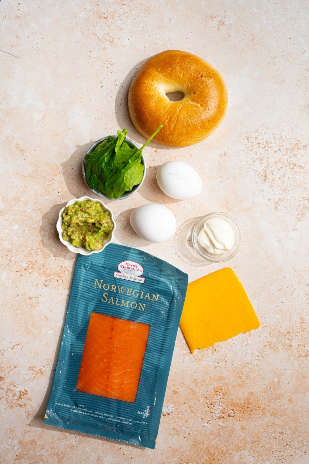 An overhead shot of ingredients to make a loaded breakfast bagel sandwich including a whole plain bagel, spinach, eggs, cheese, mashed avocado, and smoked salmon.