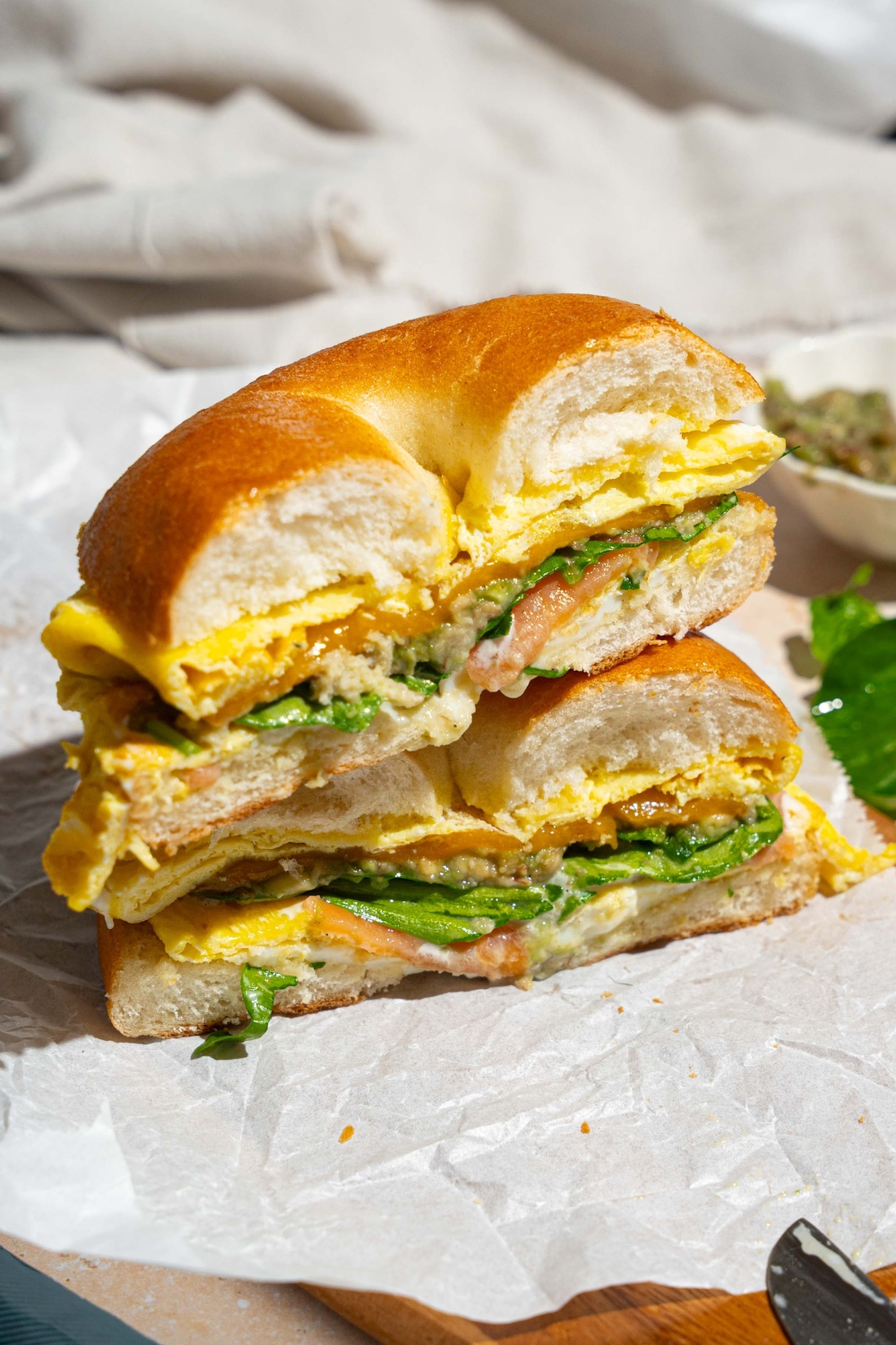 A sliced loaded bagel breakfast sandwich on a wooden board lined with parchment paper. The board is on a tan counter with a white cloth napkin.