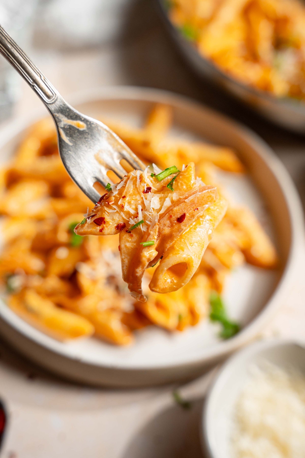 A close up of a fork with a bite of blush sauce pasta garnished with fresh parsley, shredded parmesan, and red pepper flakes. There is a plate of pasta on a tan counter blurred in the background.