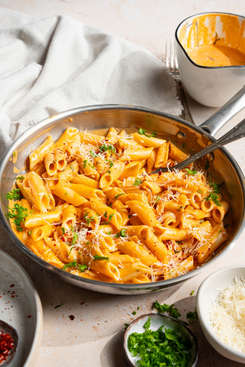 A skillet with pink sauce pasta garnished with shredded parmesan cheese, fresh parsley, and crushed red pepper. There is a fork in the pasta. The skillet is on a tan counter with a bowl of pink sauce, small plates of garnishes, and white cloth napkin.