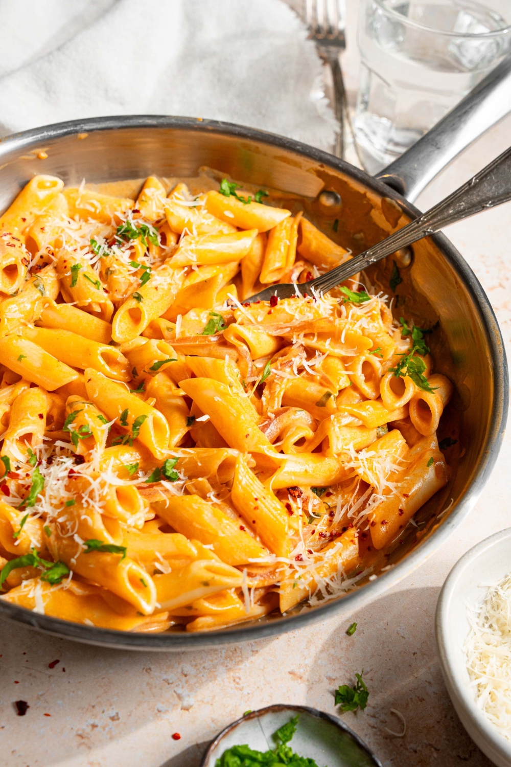 A skillet with pink sauce pasta garnished with shredded parmesan cheese, fresh parsley, and crushed red pepper. There is a fork in the pasta. The skillet is on a tan counter with small plates of garnishes, and white cloth napkin.
