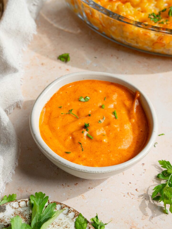 A small white bowl with campfire sauce garnished with fresh parsley. The bowl is on a tan counter with a small plate of garnishes.