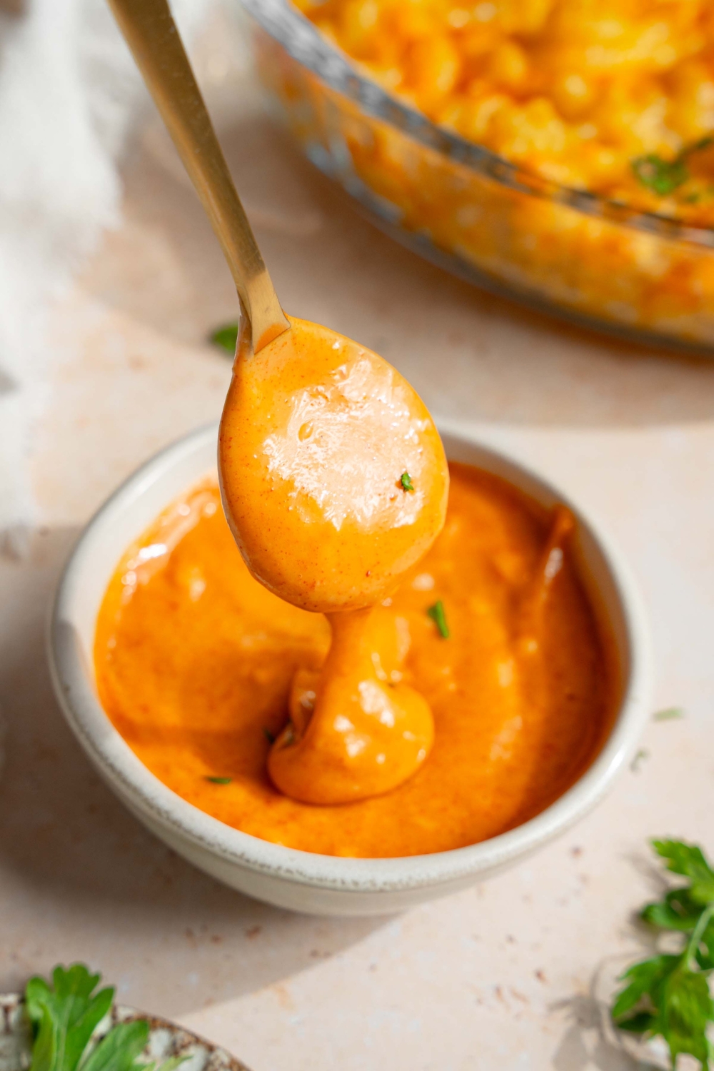 A spoon dipping in and out of a bowl of campfire sauce. The bowl is on a tan counter with a plate of garnishes.