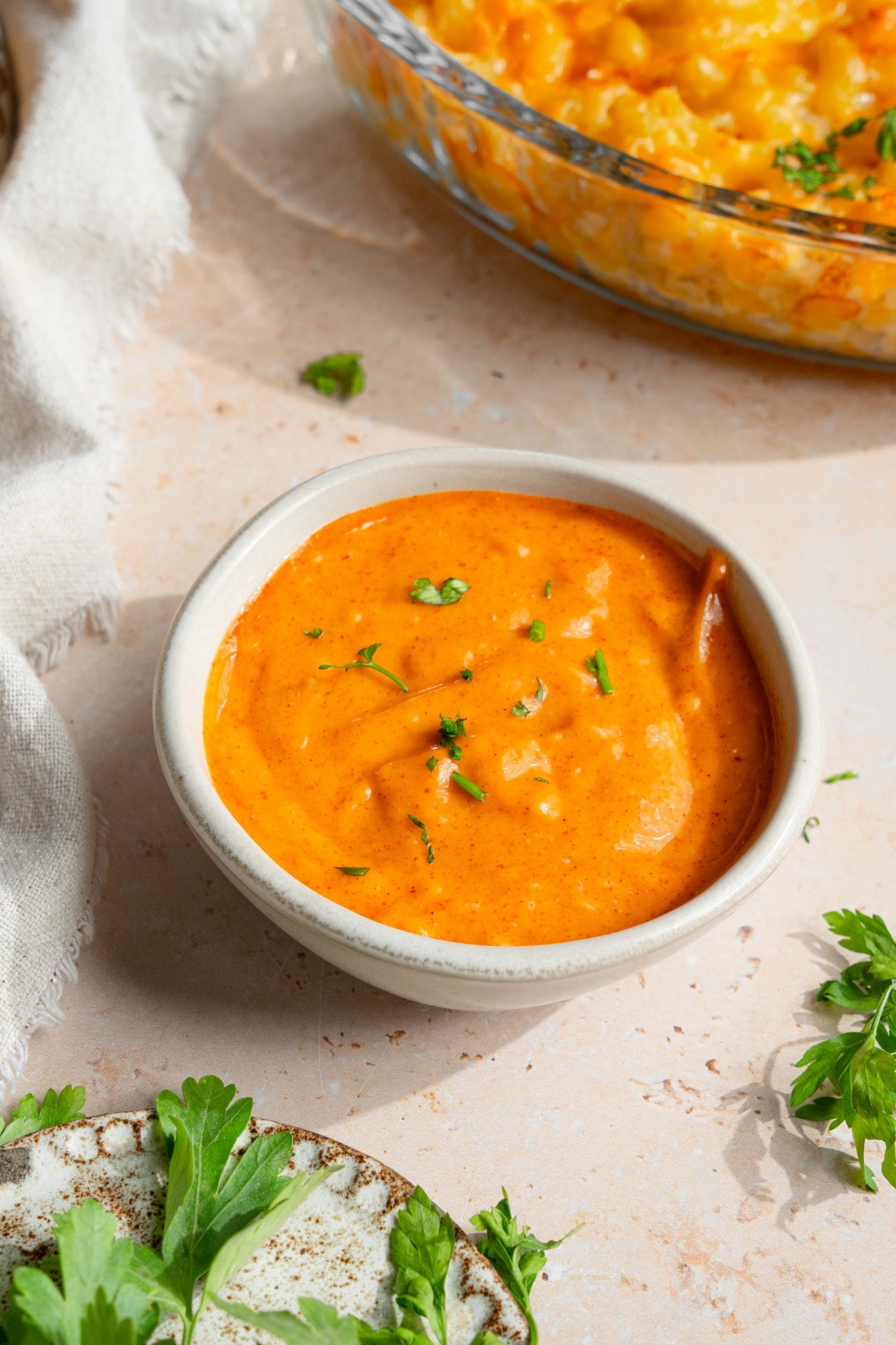A small white bowl with campfire sauce garnished with fresh parsley. The bowl is on a tan counter with a small plate of garnishes.