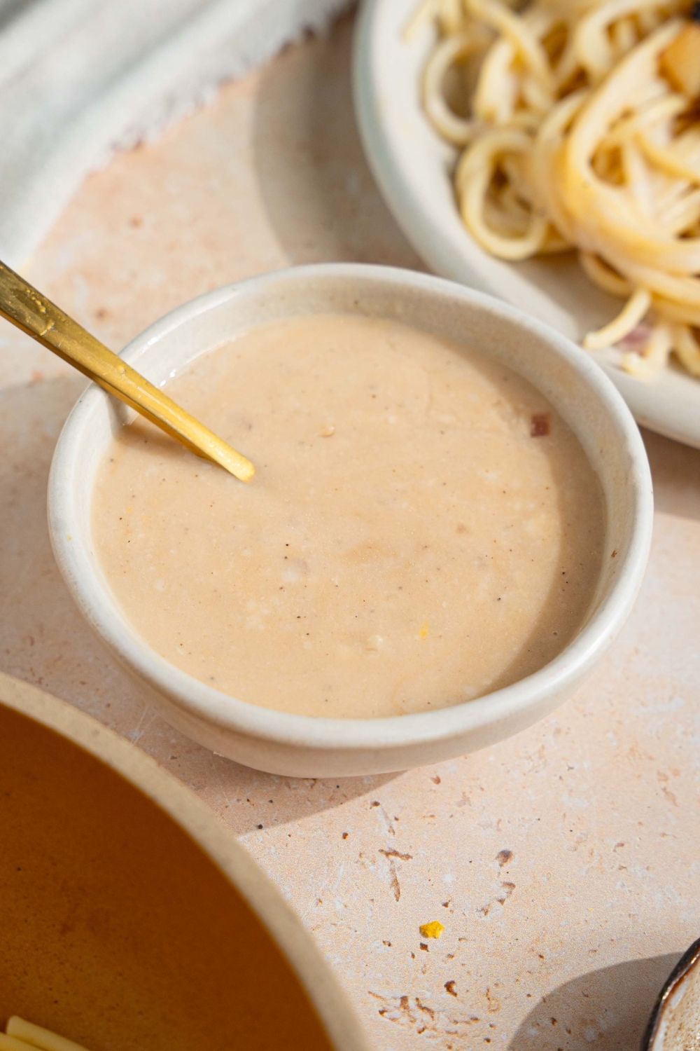 A small bowl of sherry cream sauce with a spoon in the bowl. The bowl is on a tan counter with a plate of spaghetti.