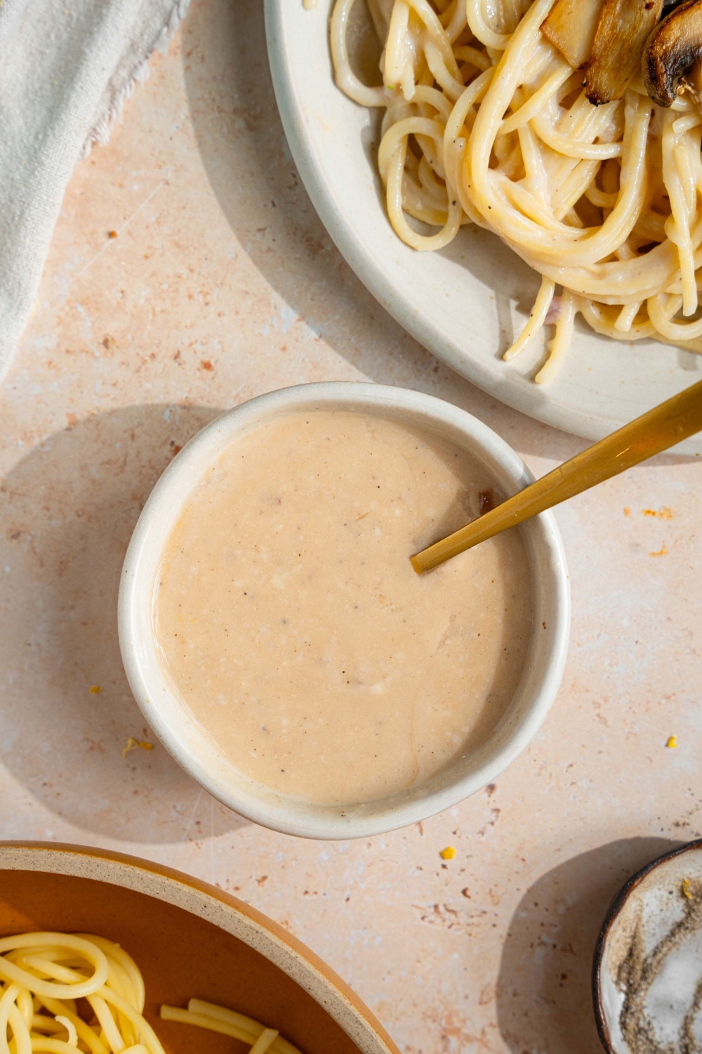 A small bowl of sherry cream sauce with a spoon in the bowl. The bowl is on a tan counter with a plate of spaghetti and sherry cream sauce.
