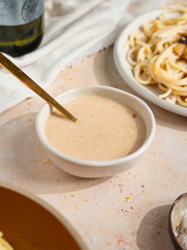 A small bowl of sherry cream sauce with a spoon in the bowl. The bowl is on a tan counter with a plate of spaghetti and white cloth napkin.