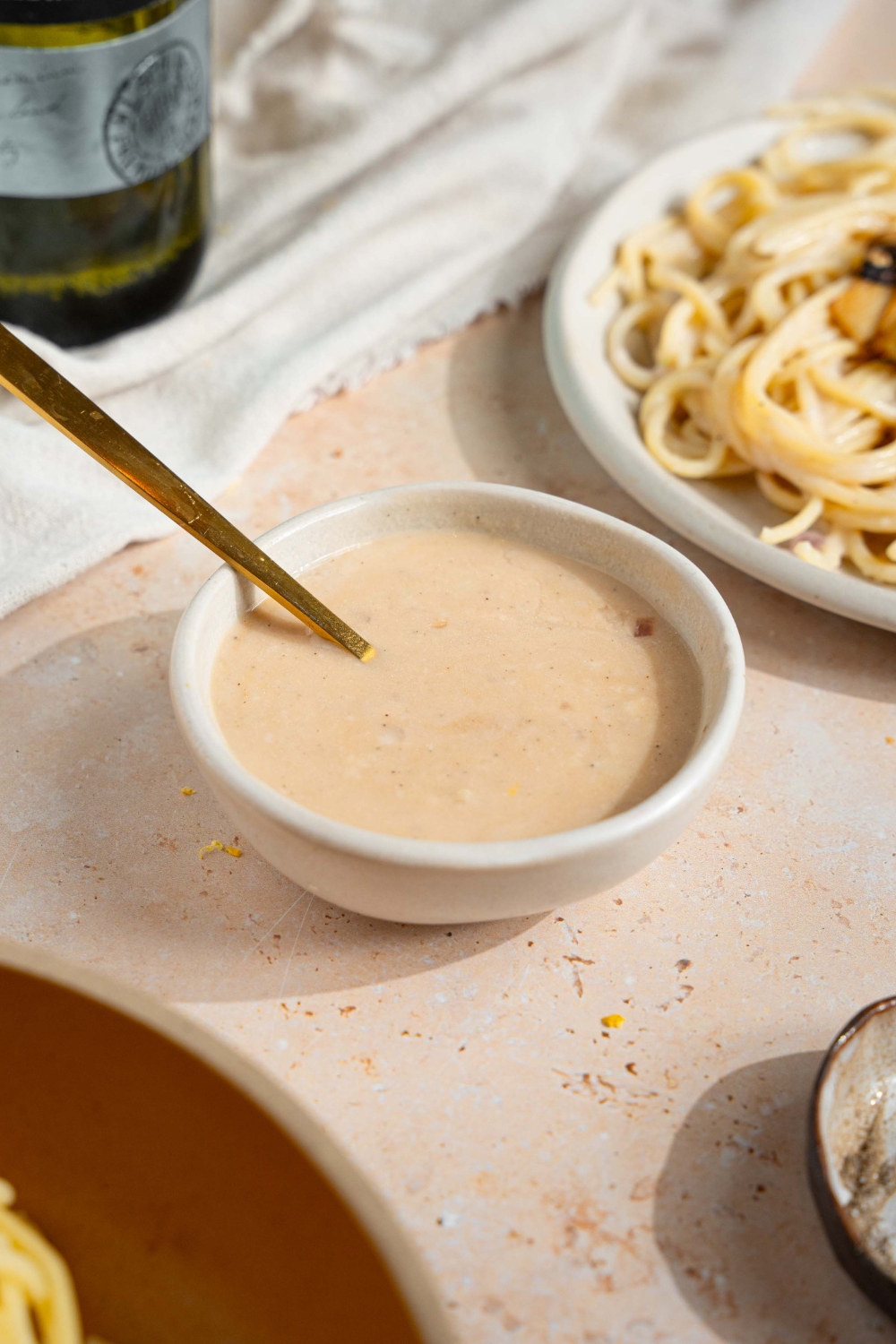 A small bowl of sherry cream sauce with a spoon in the bowl. The bowl is on a tan counter with a plate of spaghetti and white cloth napkin.