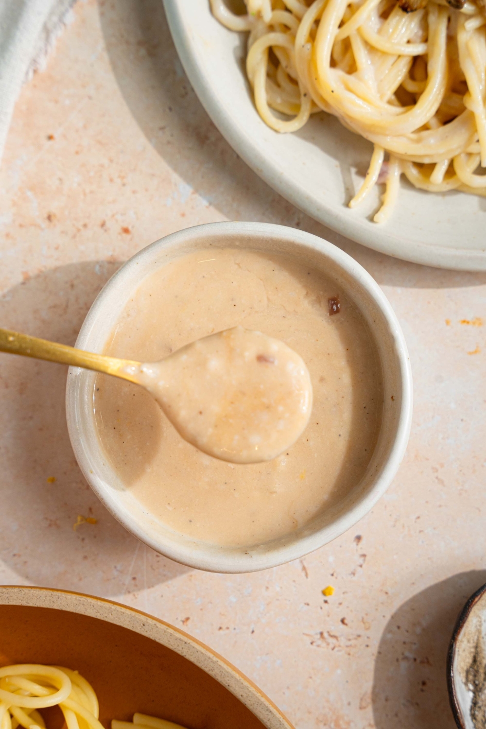 A small bowl of sherry cream sauce with a spoon dipping in the bowl. The bowl is on a tan counter with a plate of spaghetti.
