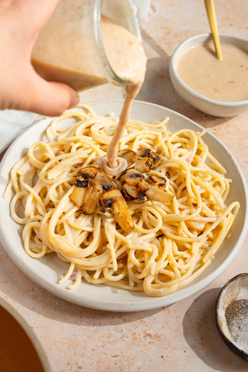 A white plate with spaghetti topped with mushrooms. A hand is pouring a jar of sherry cream sauce over the dish. The plate is on a tan counter with a bowl of cream sauce.