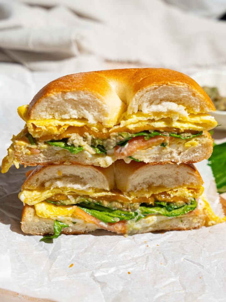 A sliced loaded bagel breakfast sandwich on a wooden board lined with parchment paper. The board is on a tan counter with a white cloth napkin.