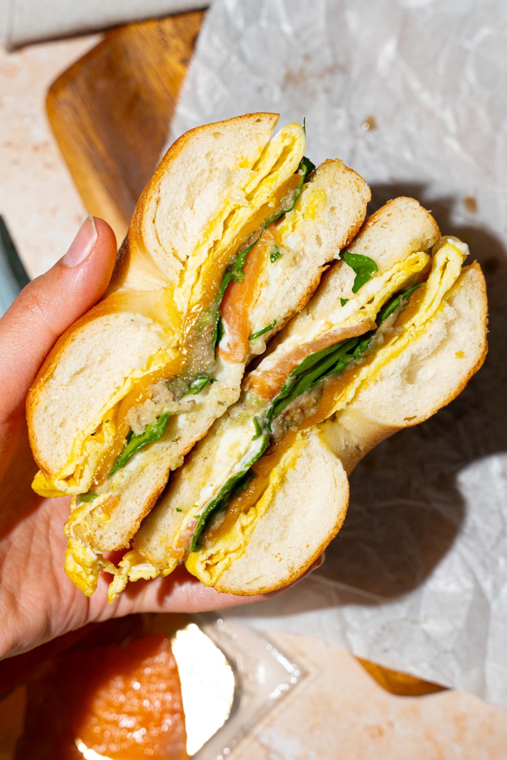 A hand holing a sliced loaded bagel breakfast sandwich. There is a wooden board lined with parchment paper in the background.