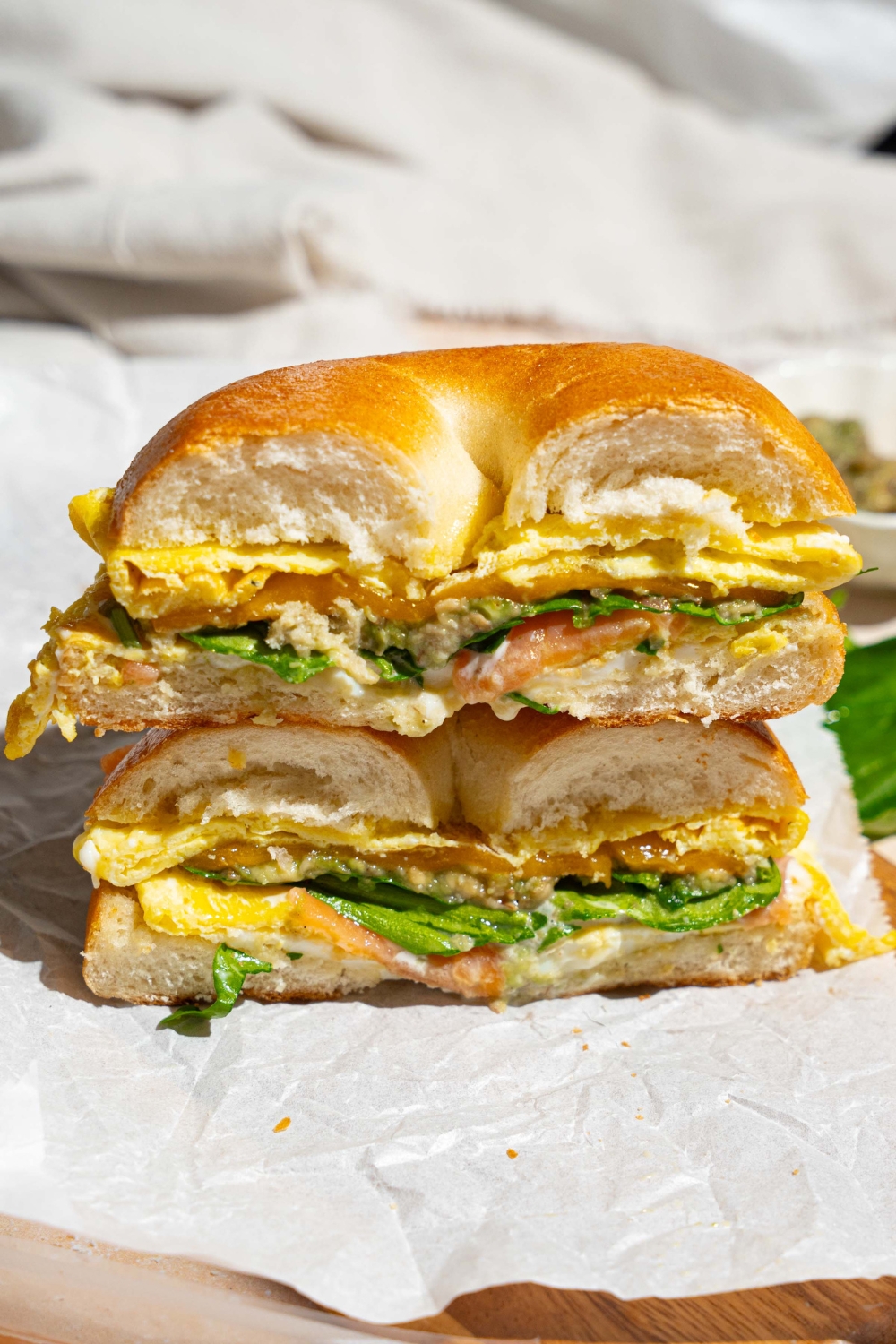 A sliced loaded bagel breakfast sandwich on a wooden board lined with parchment paper. The board is on a tan counter with a white cloth napkin.