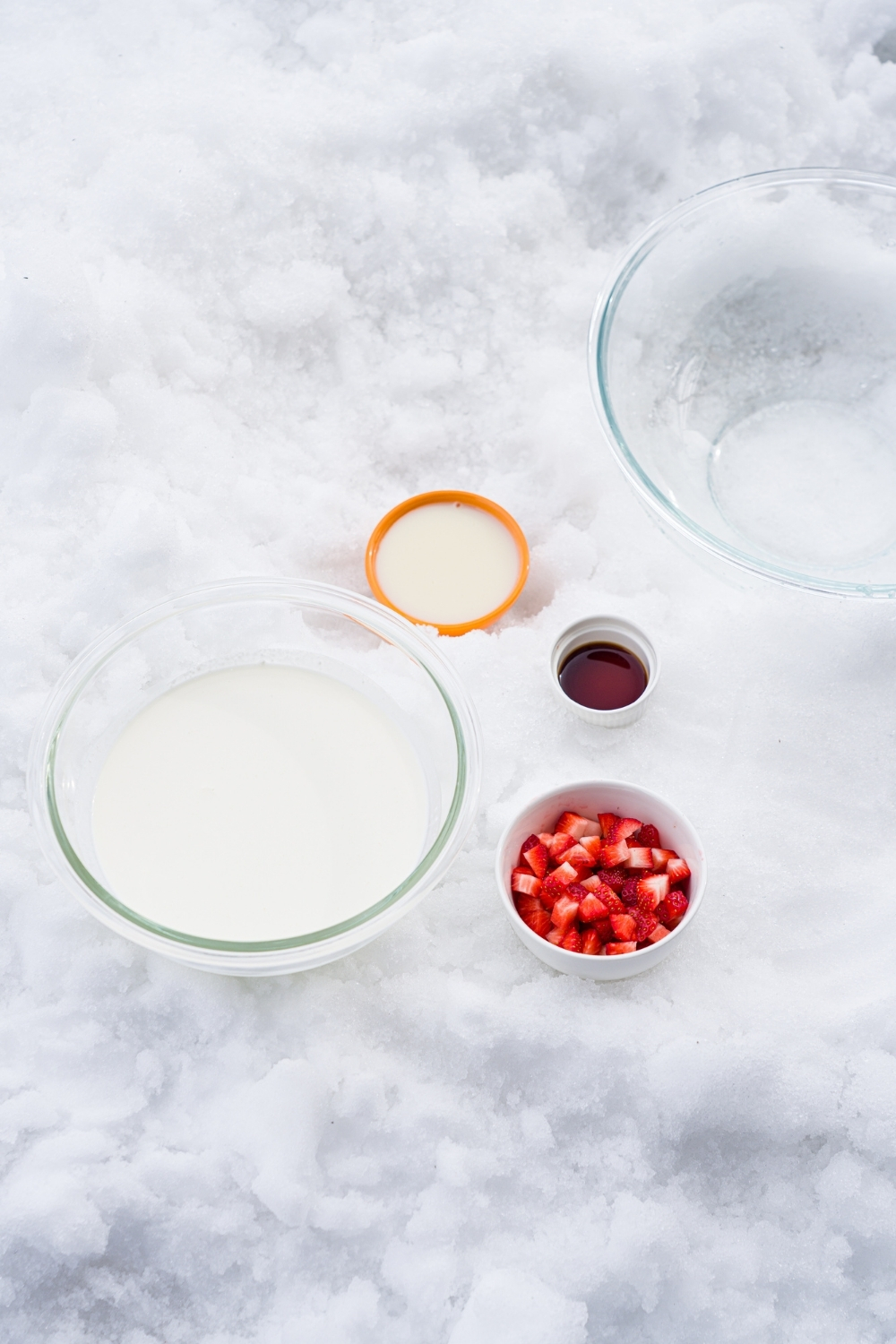 A pile of snow with ingredients to make ice cream including heavy cream, sweetened condensed milk, strawberries, and vanilla.