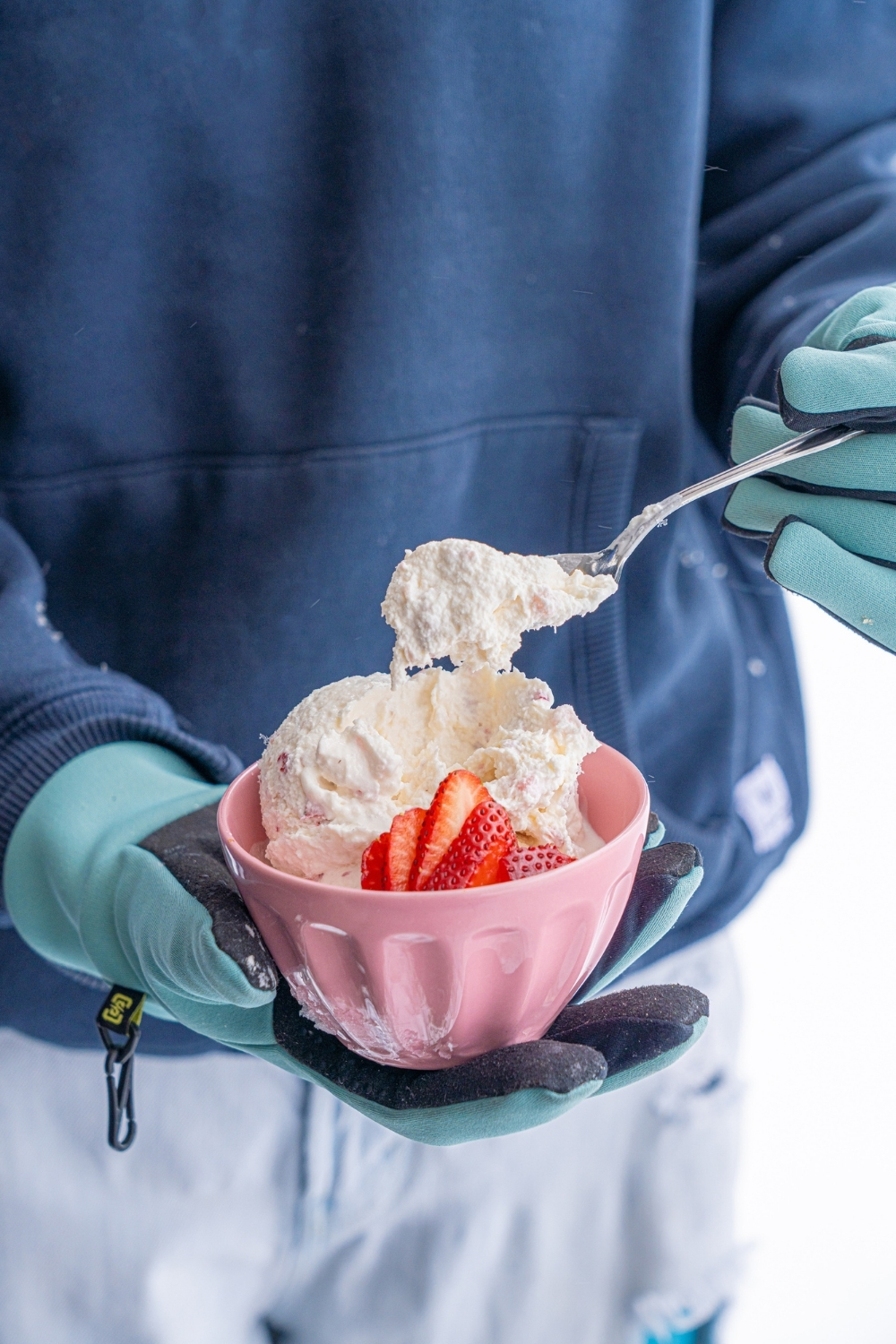 A pink bowl with scoops of snow ice cream with sliced strawberries. A hand is dipping a spoon into the ice cream.