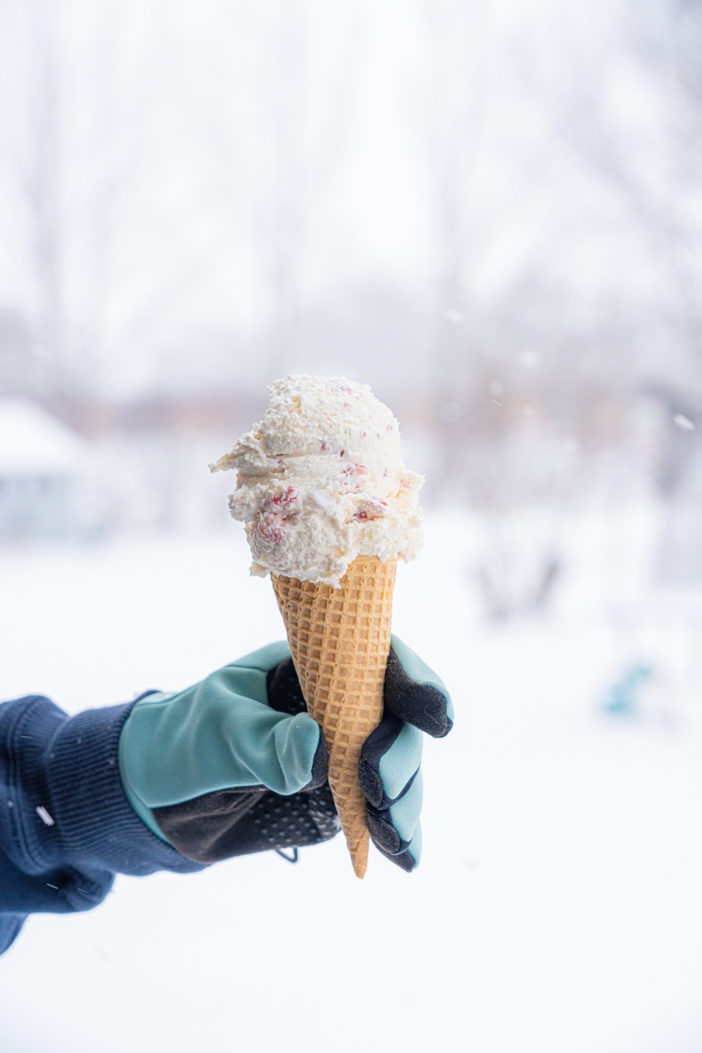 A hand holding an ice cream cone of snow ice cream with strawberries. There is a snowy field in the background.