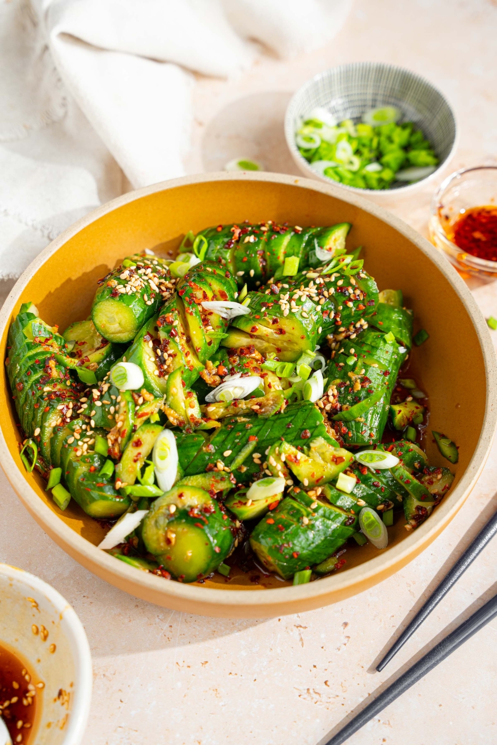 A bowl of spicy Asian cucumber salad tossed in dressing and garnished with slice green onions, crushed red pepper, and sesame seeds. The bowl is on a tan counter with a small bowl of sliced green onion, a pair of chopsticks, and a white cloth napkin.