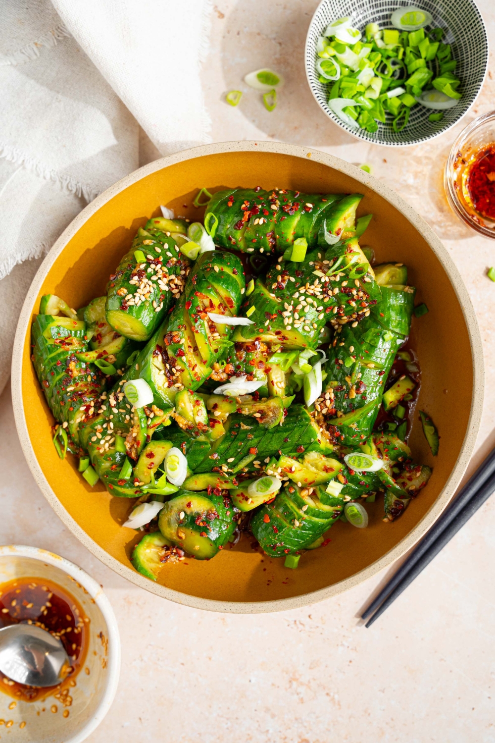 A bowl of spicy Asian cucumber salad tossed in dressing and garnished with slice green onions, crushed red pepper, and sesame seeds. The bowl is on a tan counter with a small bowl of sliced green onion, a pair of chopsticks, and a white cloth napkin.