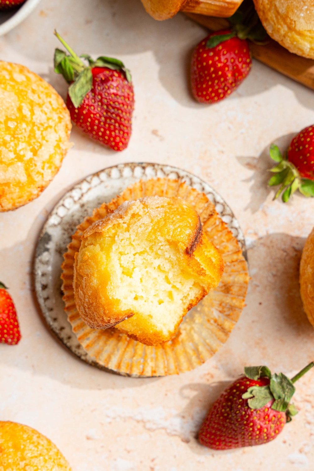 A small plate with a vanilla muffin with the liner unwrapped. A bite is taken from the muffin. The plate is on a tan counter with strawberries and additional muffins.