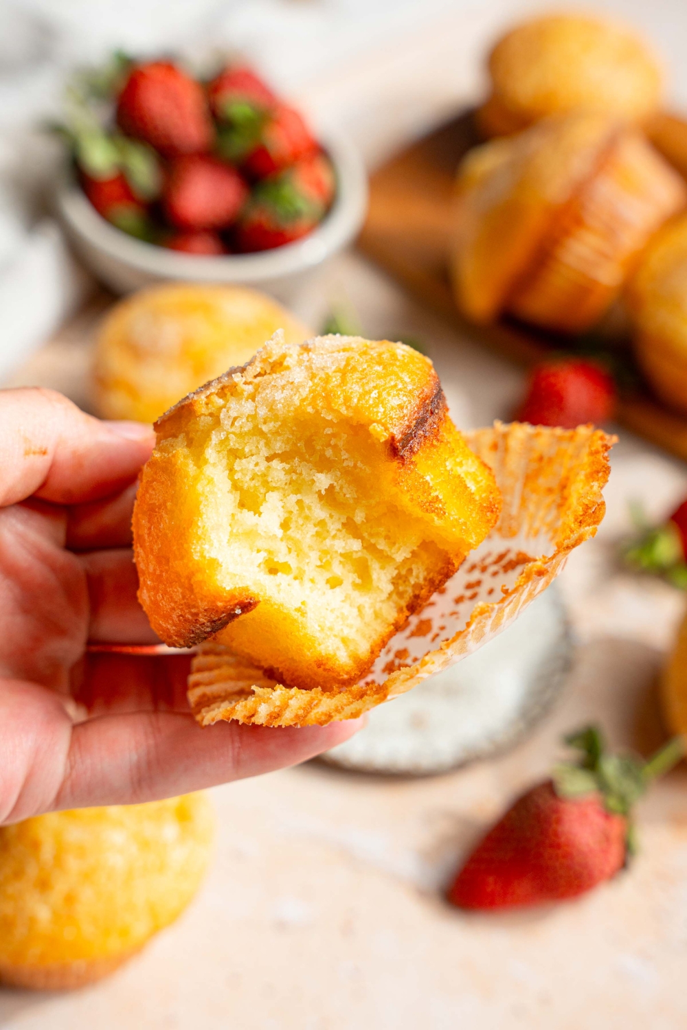 A close up of vanilla muffin with the lining unwrapped and a bite taken from the muffin. There is a tan counter with a wooden board with muffins and strawberries blurred in the background.