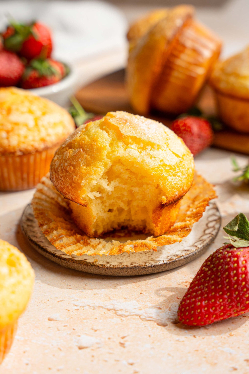 A small plate with a vanilla muffin with the liner unwrapped. A bite is taken from the muffin. The plate is on a tan counter with a wooden board of muffins and bowl of strawberries.