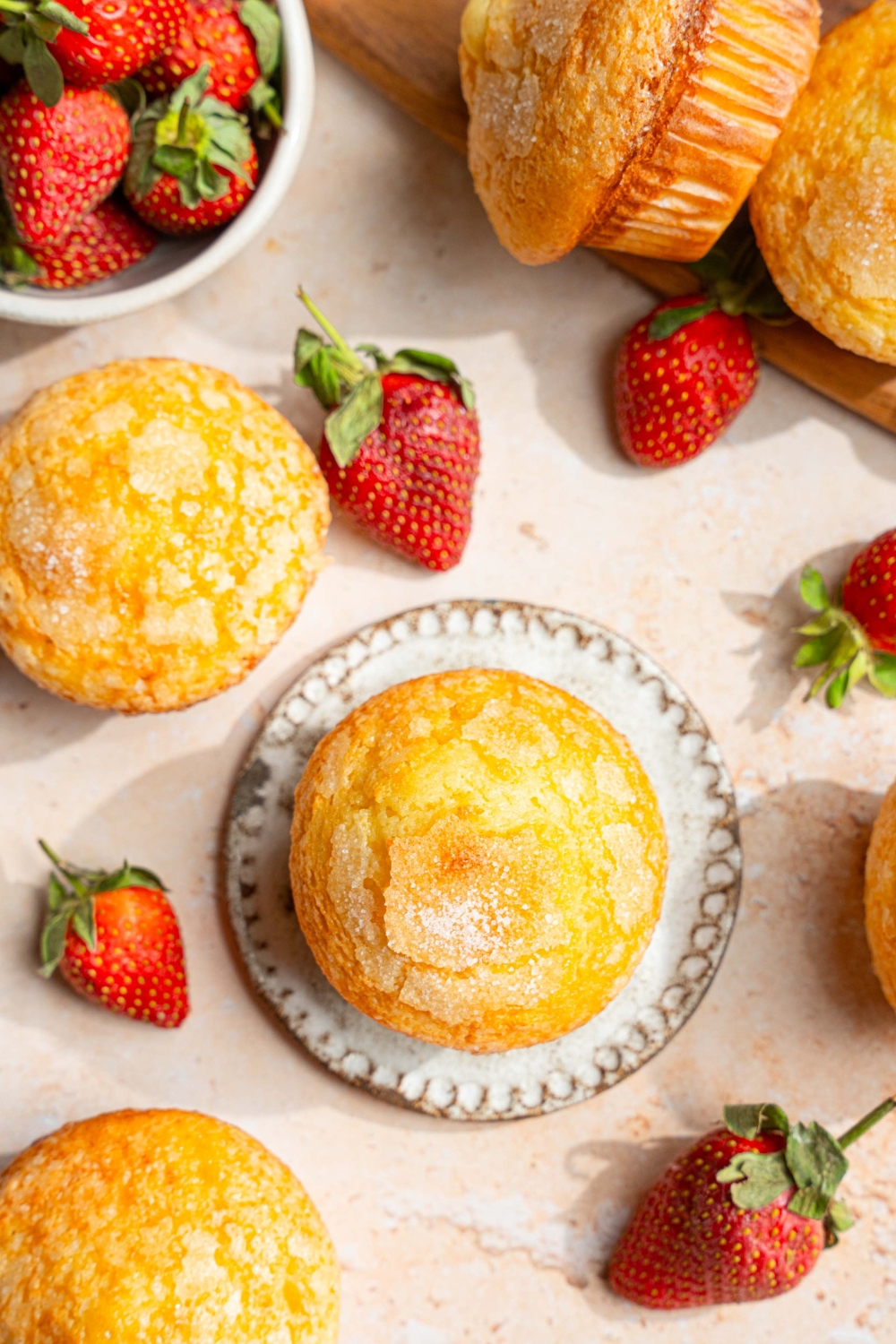 A vanilla muffin on a small plate. The plate is on a tan counter with a wooden board of muffins and a bowl of strawberries.