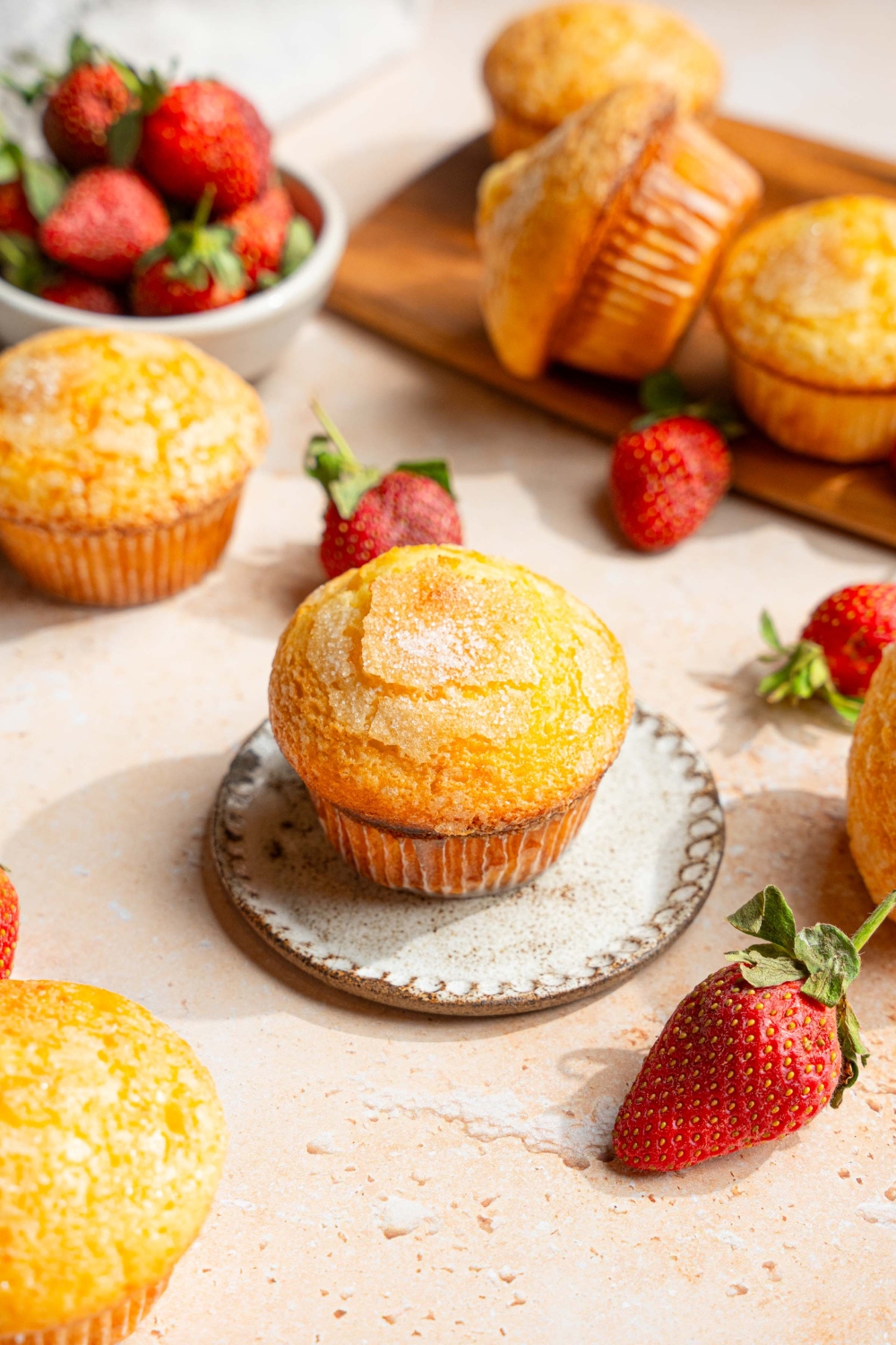 A vanilla muffin on a small plate. The plate is on a tan counter with a wooden board of muffins and a bowl of strawberries.