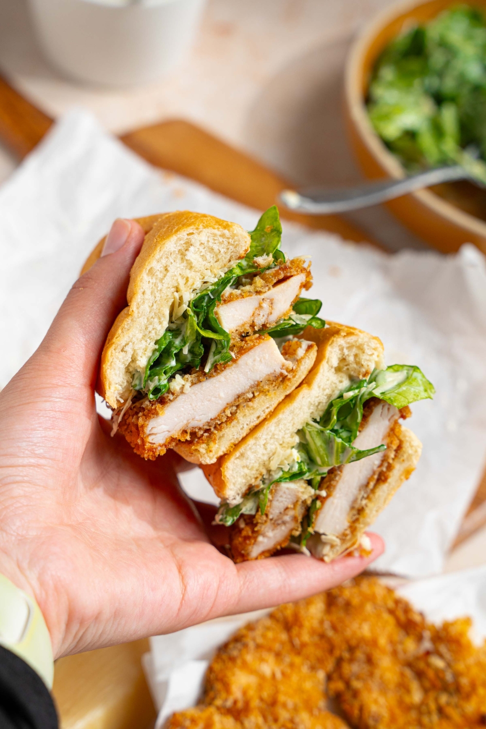 A hand holding a sliced chicken caesar sandwich with a wooden board lined with parchment paper blurred in the background.