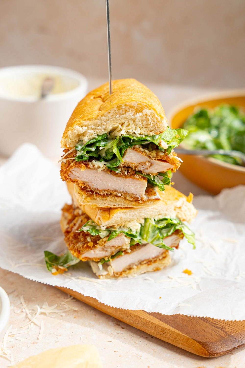 A sliced chicken caesar sandwich stacked and skewered. The sandwich is on a wooden board lined with parchment paper. The board is on a tan counter with a bowl of caesar salad.