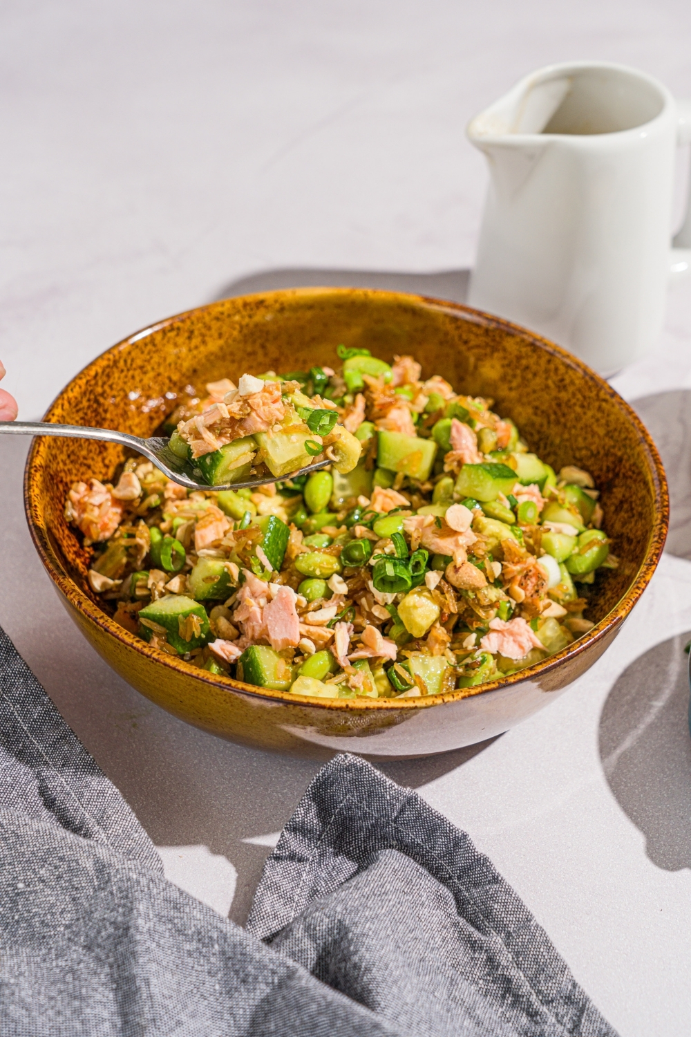 A bowl of crispy rice salmon salad garnished with sliced green onion. There is a fork with a bite of salad. The bowl is on a marble counter with a gray cloth napkin and saucer of dressing.