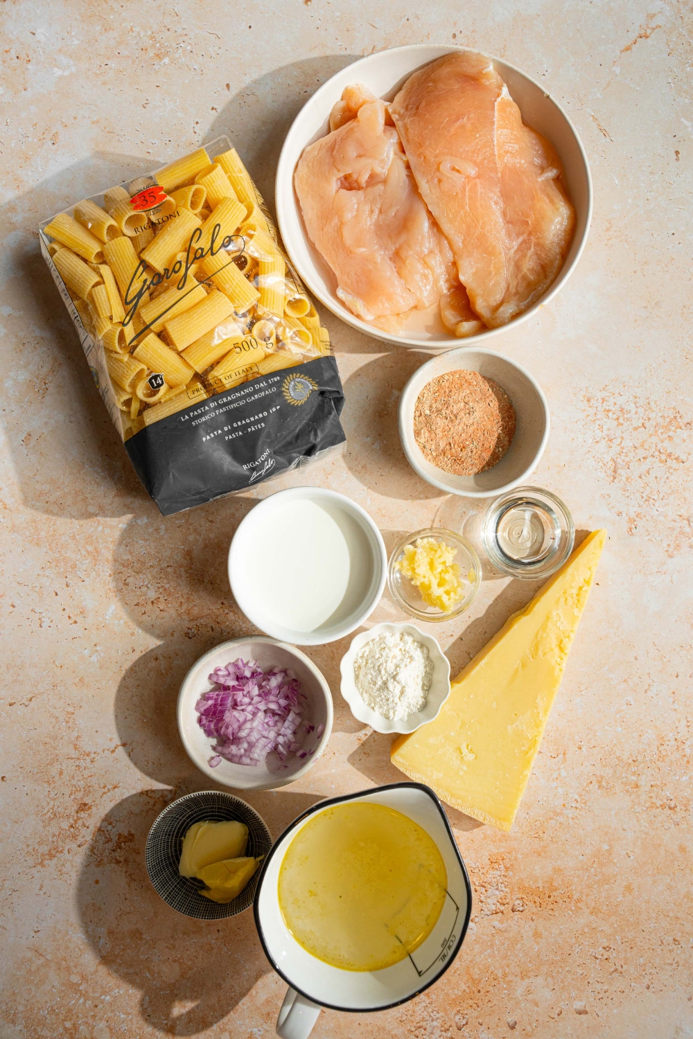 An overhead shot of several bowls in various sizes containing ingredients to make garlic parmesan chicken pasta including chicken breasts, rigatoni pasta, heavy cream, chicken broth, onion, garlic, flour, butter, oil, and parmesan cheese.