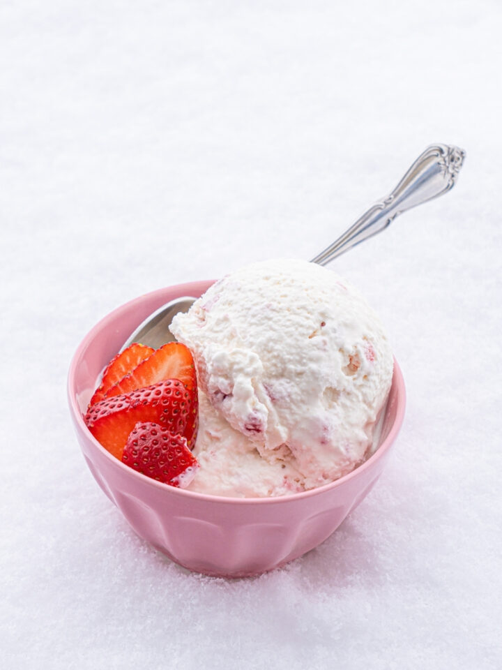 A pink bowl with scoops of snow ice cream with sliced strawberries with a spoon in the bowl. The bowl is sitting in a pile of snow.