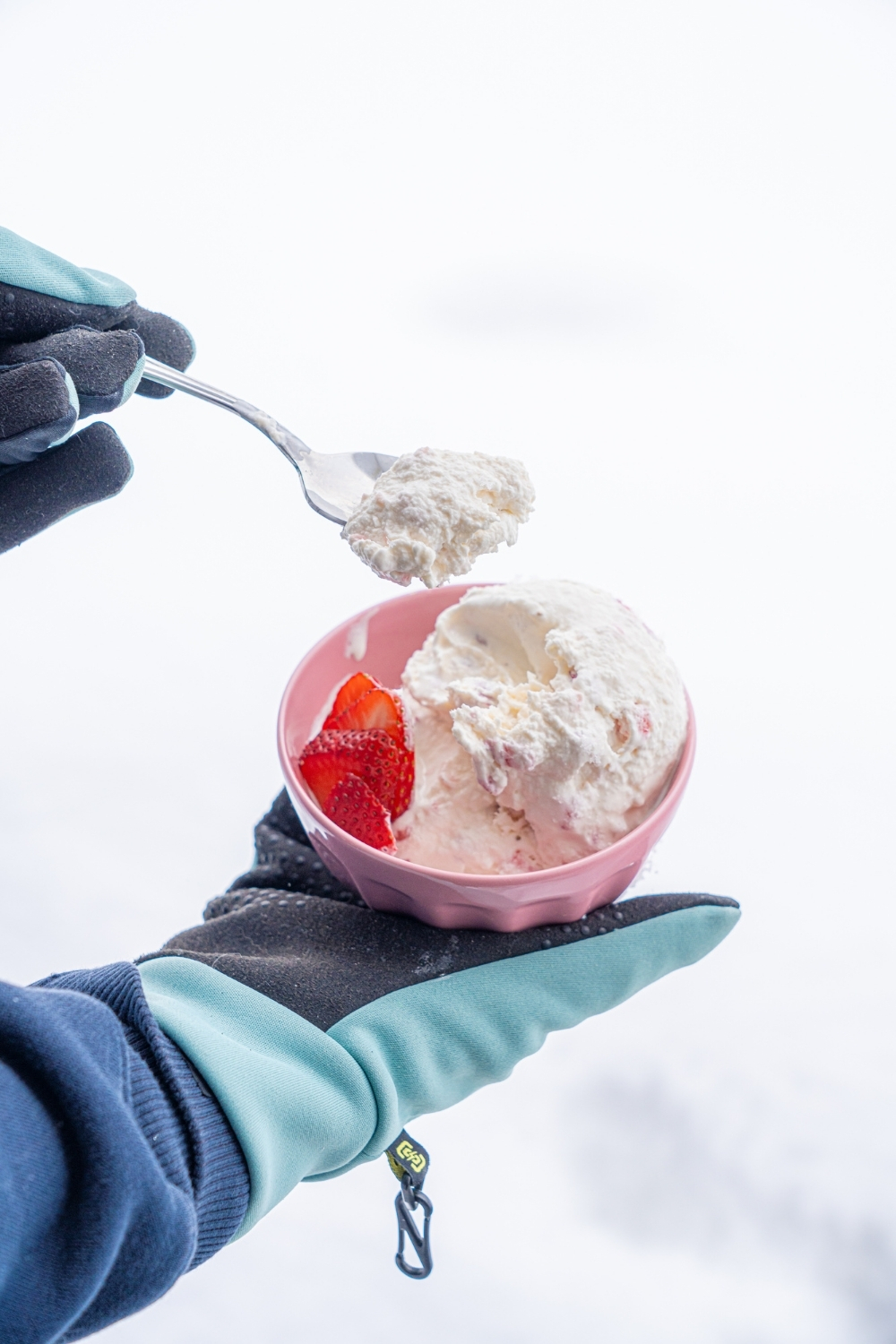 A pink bowl with scoops of snow ice cream with sliced strawberries. A hand is dipping a spoon into the ice cream.