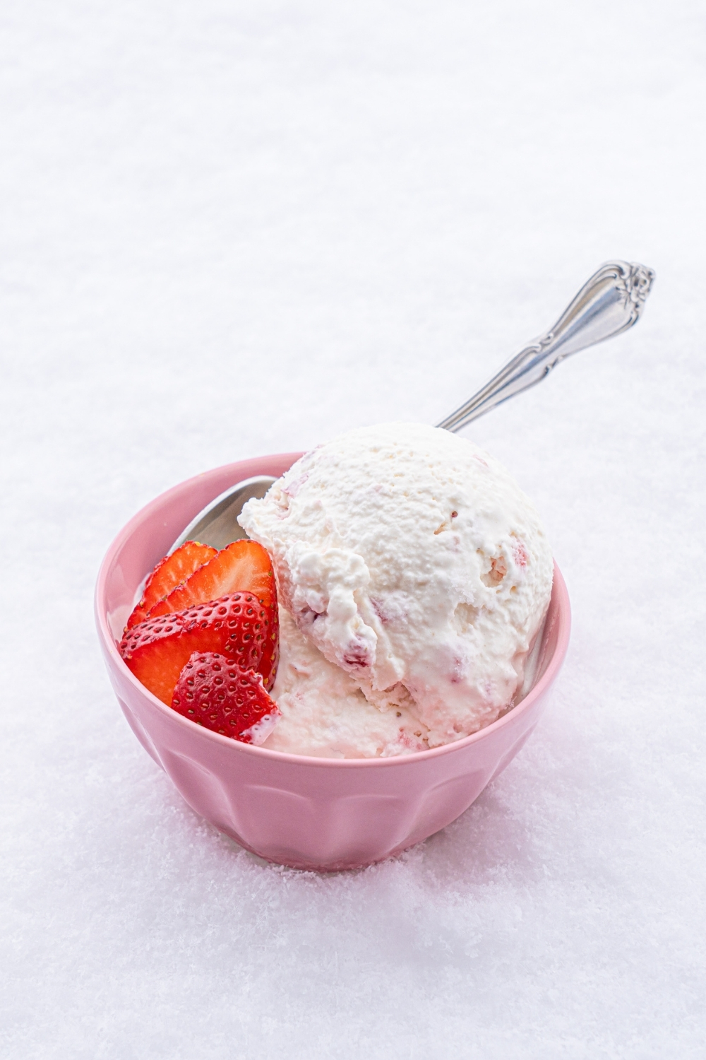 A pink bowl with scoops of snow ice cream with sliced strawberries with a spoon in the bowl. The bowl is sitting in a pile of snow.