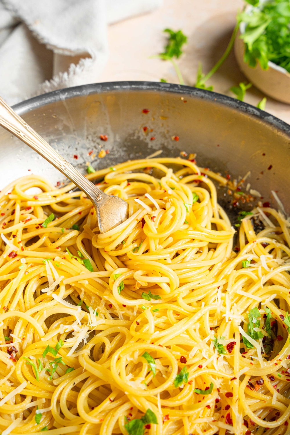 A skillet with spaghetti tossed in white wine sauce garnished with fresh parsley and crushed red pepper. A fork is twirling a bite of pasta. The skillet is on a tan counter with a white cloth napkin and small bowl of parsley.