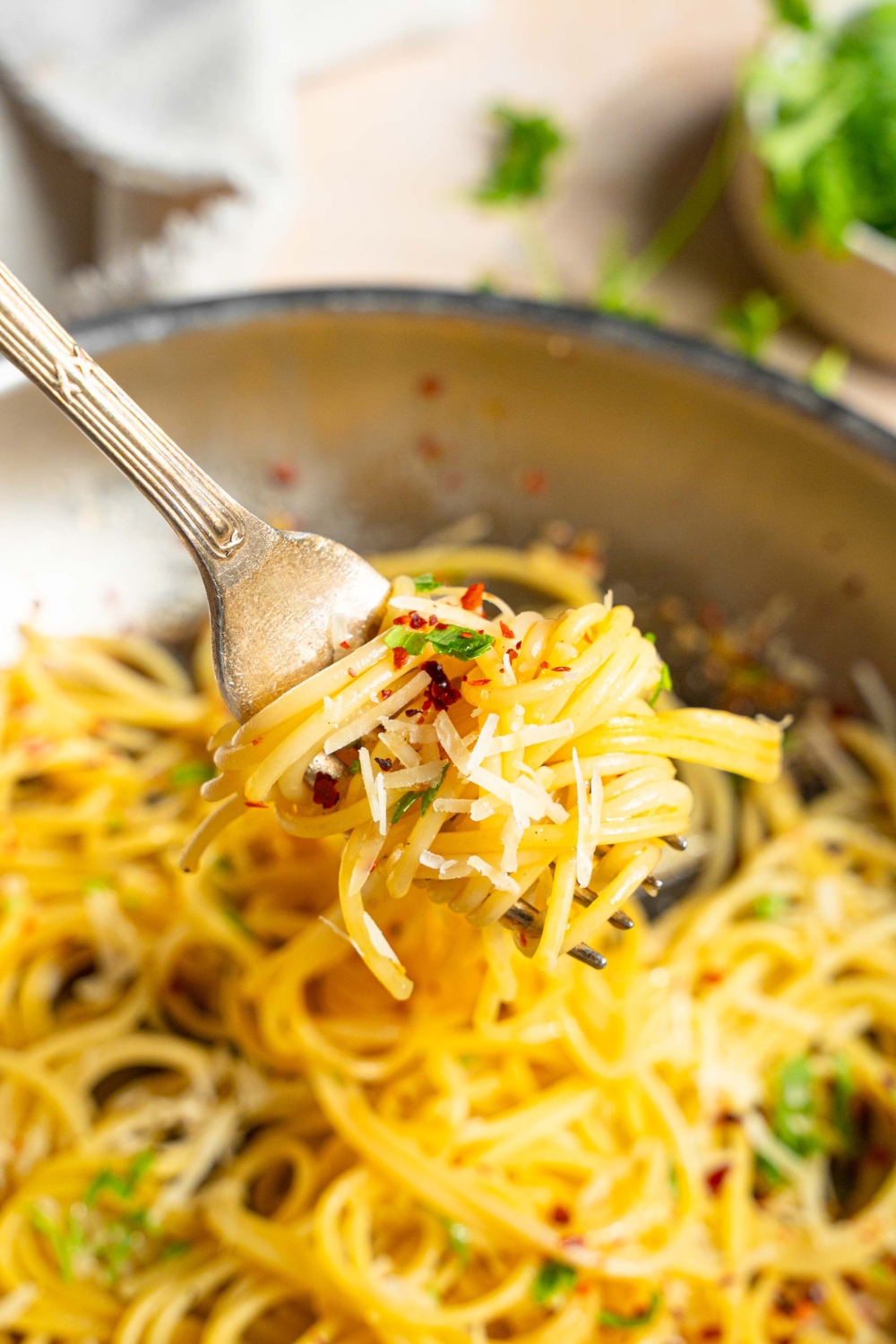 A close up of a fork with a bite of spaghetti in white wine sauce garnished with fresh parsley, crushed red pepper, and cheese. There is a skillet of spaghetti blurred in the background.