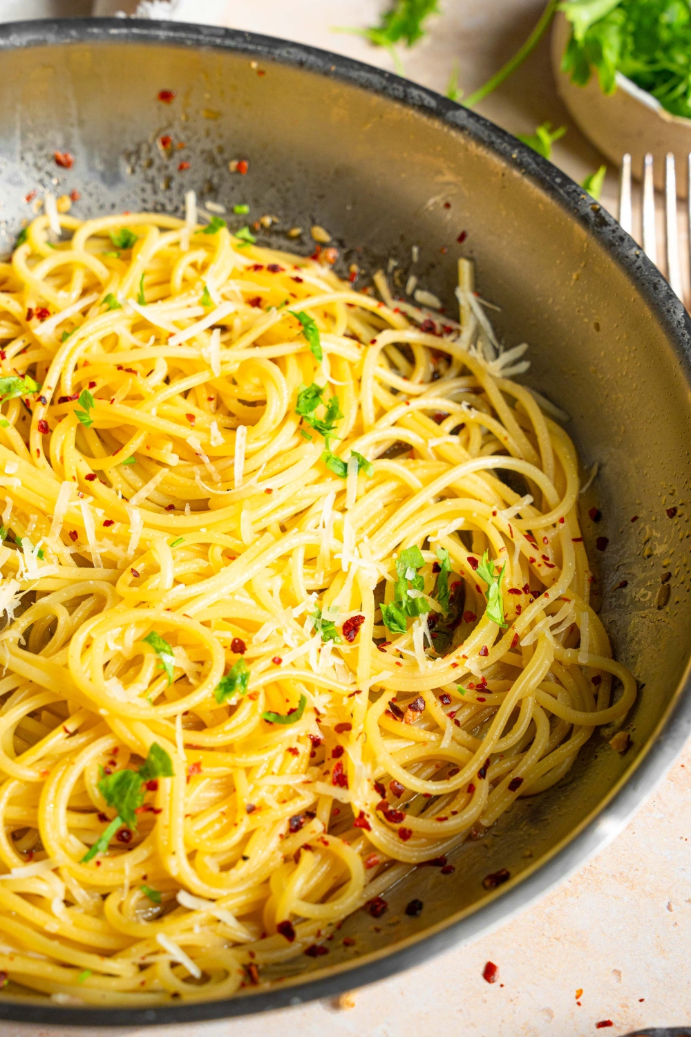A skillet with spaghetti tossed in white wine sauce garnished with fresh parsley and crushed red pepper. The skillet is on a tan counter with a white cloth napkin and small bowl of parsley.