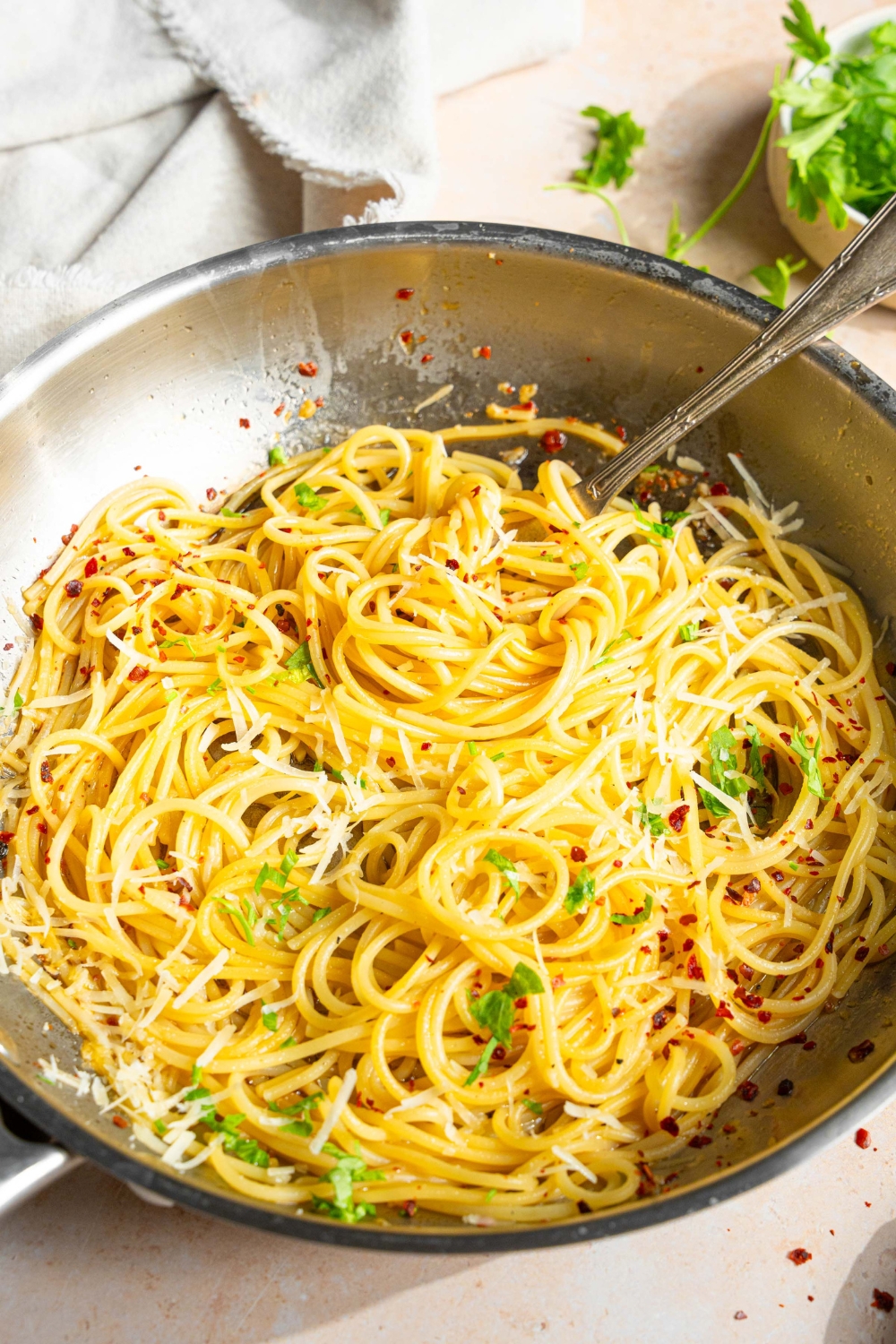 A skillet with spaghetti tossed in white wine sauce garnished with fresh parsley and crushed red pepper. A fork is in the pasta. The skillet is on a tan counter with a white cloth napkin and small bowl of parsley.
