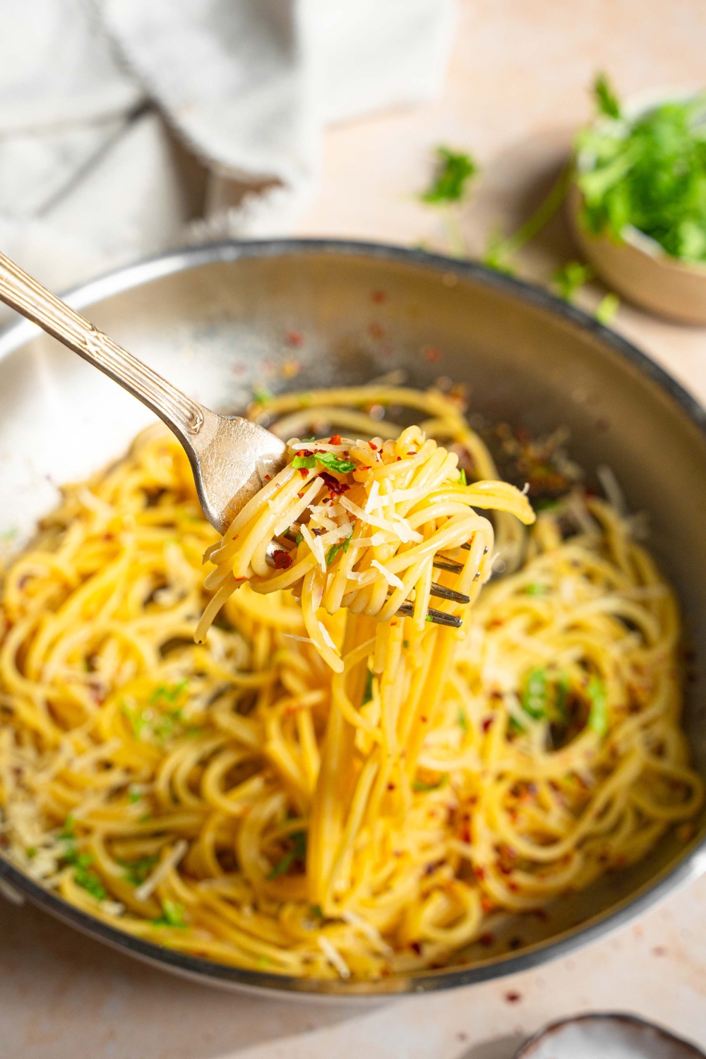 A close up of a fork with a bite of spaghetti in white wine sauce garnished with fresh parsley, crushed red pepper, and cheese. There is a skillet of spaghetti blurred in the background.