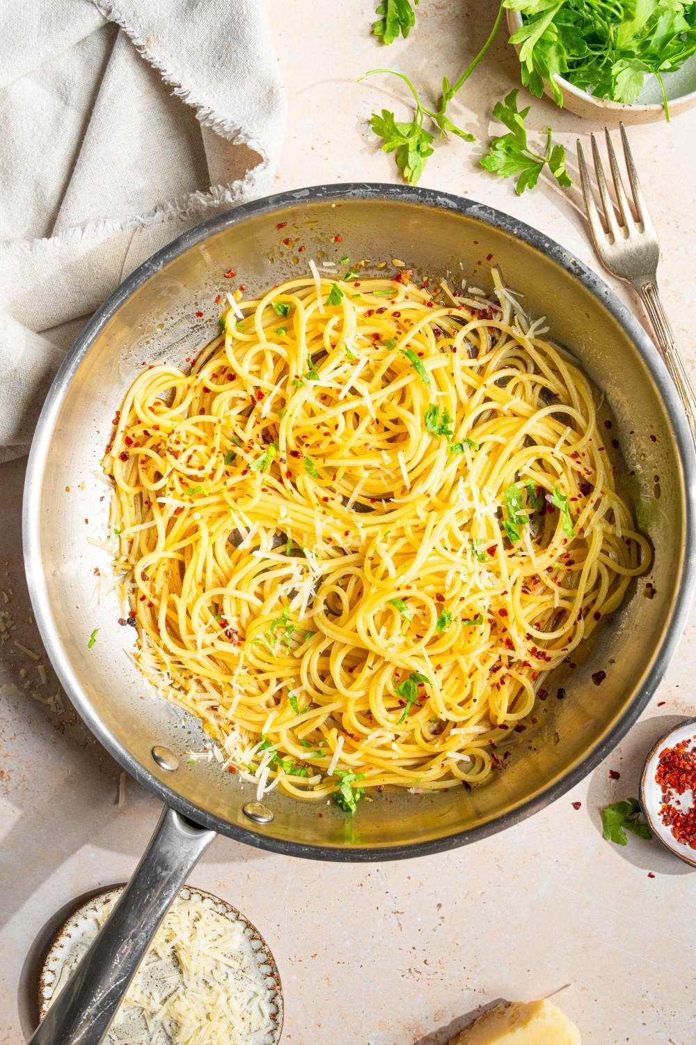 A skillet with spaghetti tossed in white wine sauce garnished with fresh parsley and crushed red pepper. The skillet is on a tan counter with a white cloth napkin and small bowl of parsley.