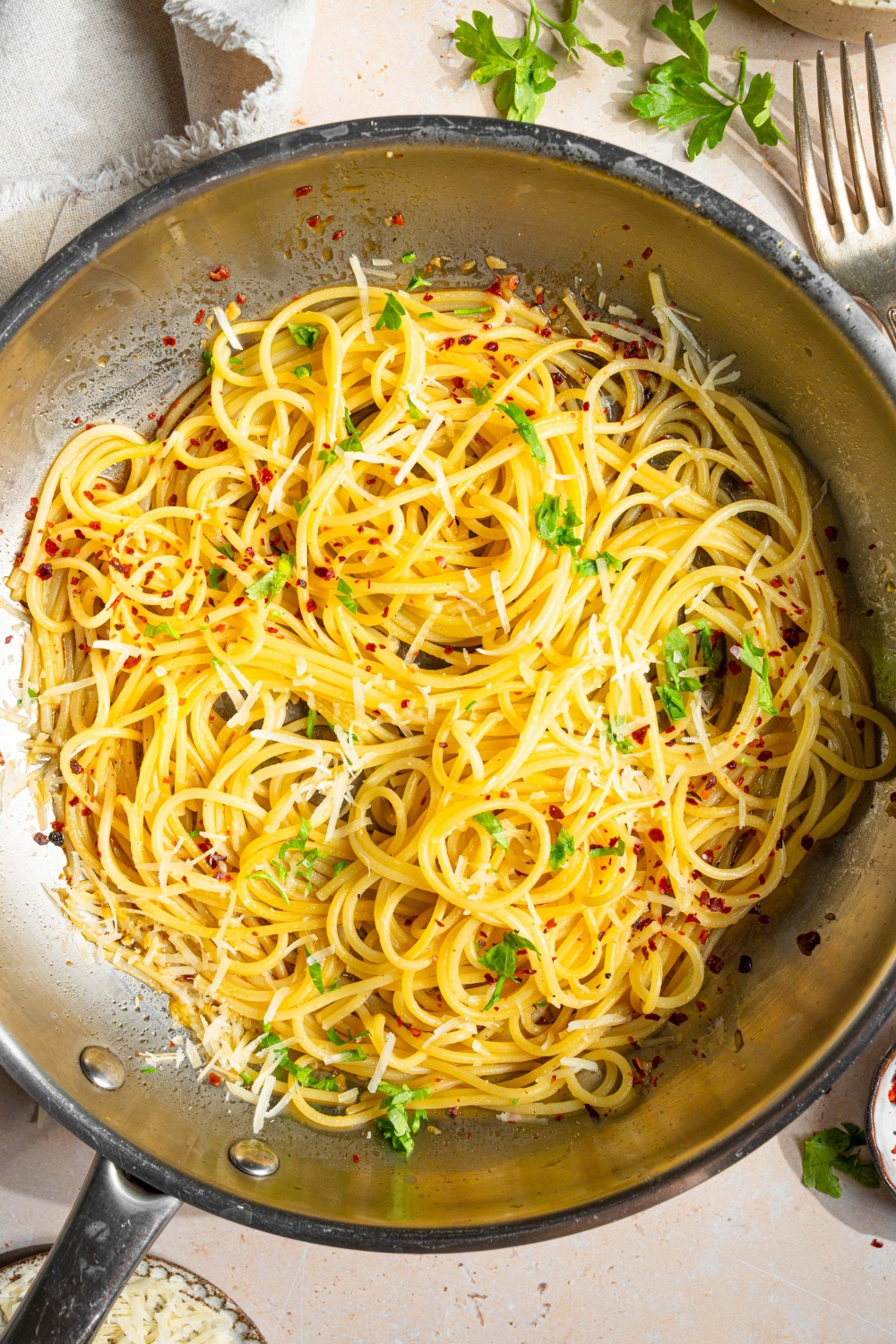 A skillet with spaghetti tossed in white wine sauce garnished with fresh parsley and crushed red pepper. The skillet is on a tan counter with a white cloth napkin and small bowl of parsley.