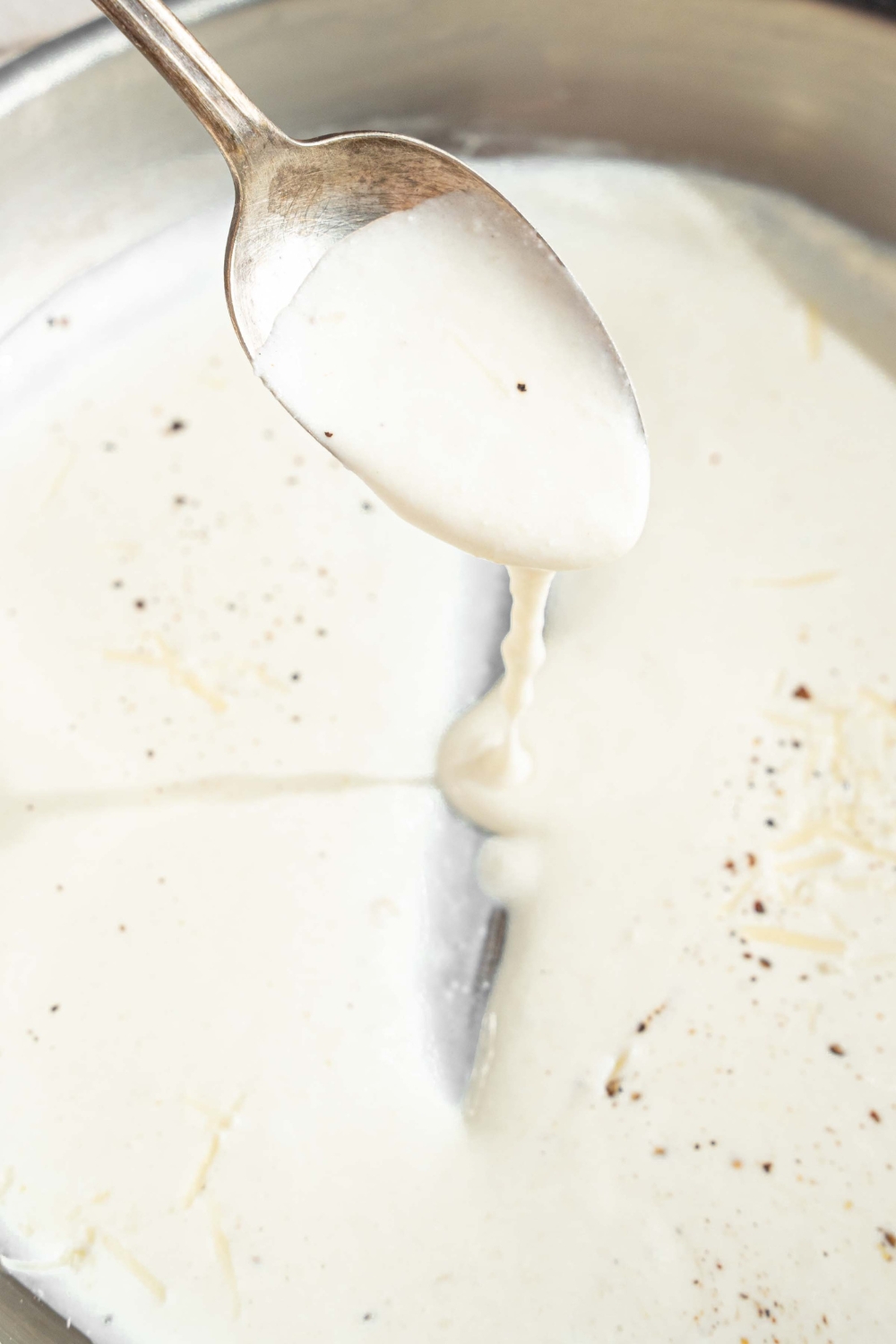 A skillet with alfredo sauce with cream cheese finished with cracked black pepper. A spoon is stirring the sauce.