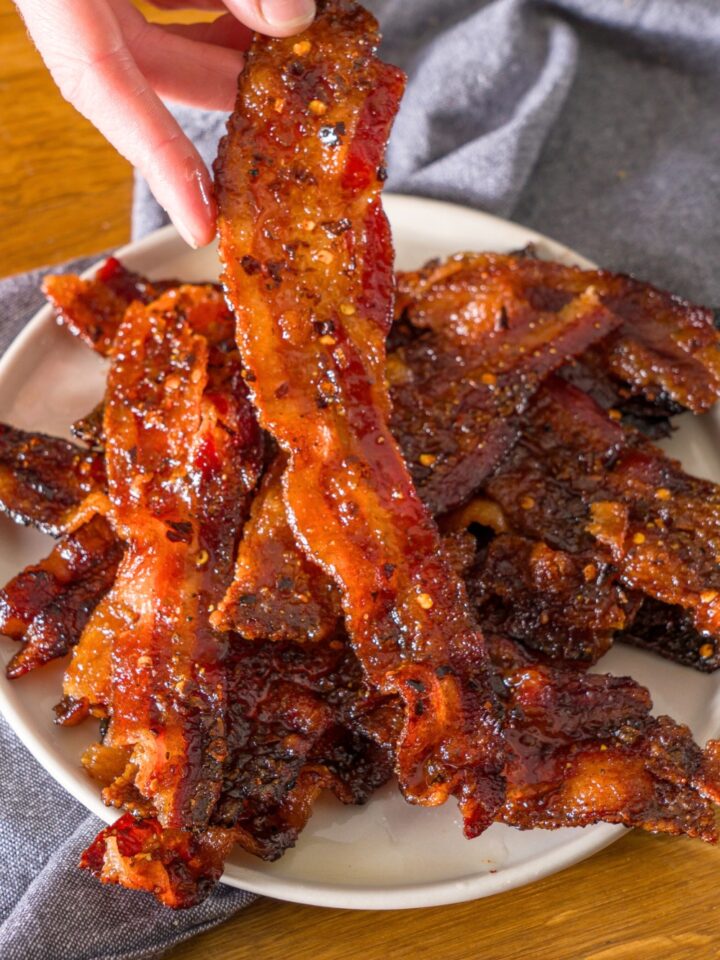 A hand holding a strip of candied bacon in front of a plate with a bunch of pieces of the bacon on it.