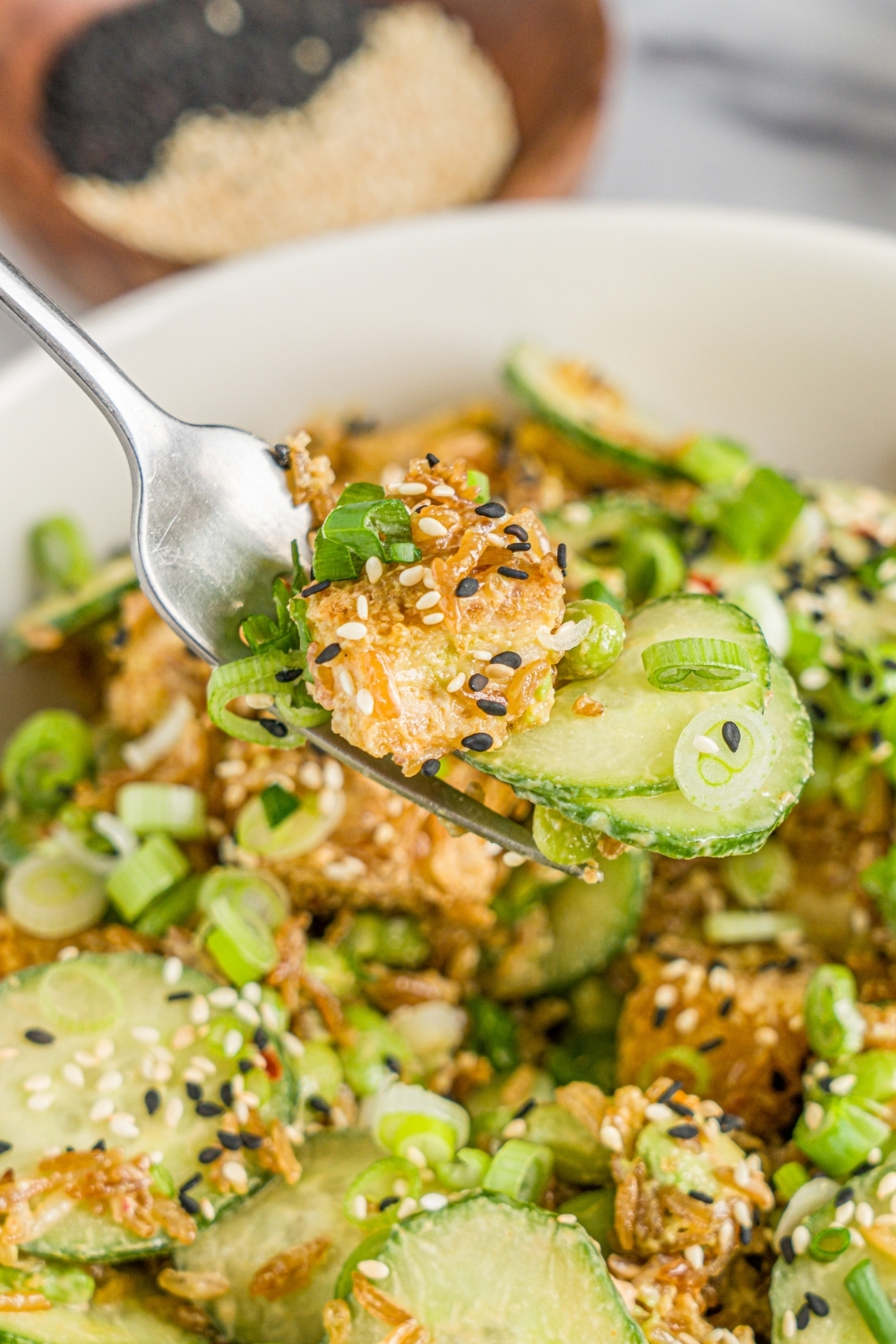 A white bowl with crispy rice chicken and cucumber salad garnished with sliced green onion and sesame seeds. There is a fork with a bite of salad over the bowl.