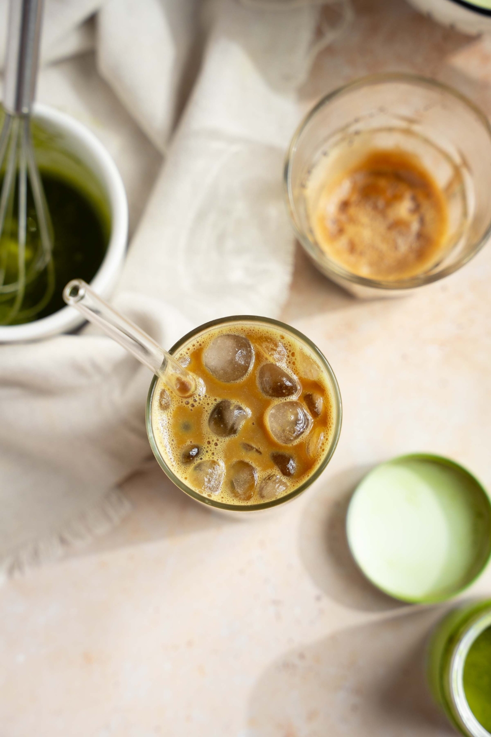 An overhead shot of an iced dirty matcha latte with a glass straw on a tan counter with a small bowl and whisk of matcha, small tin of matcha powder, and glass of poured espresso.