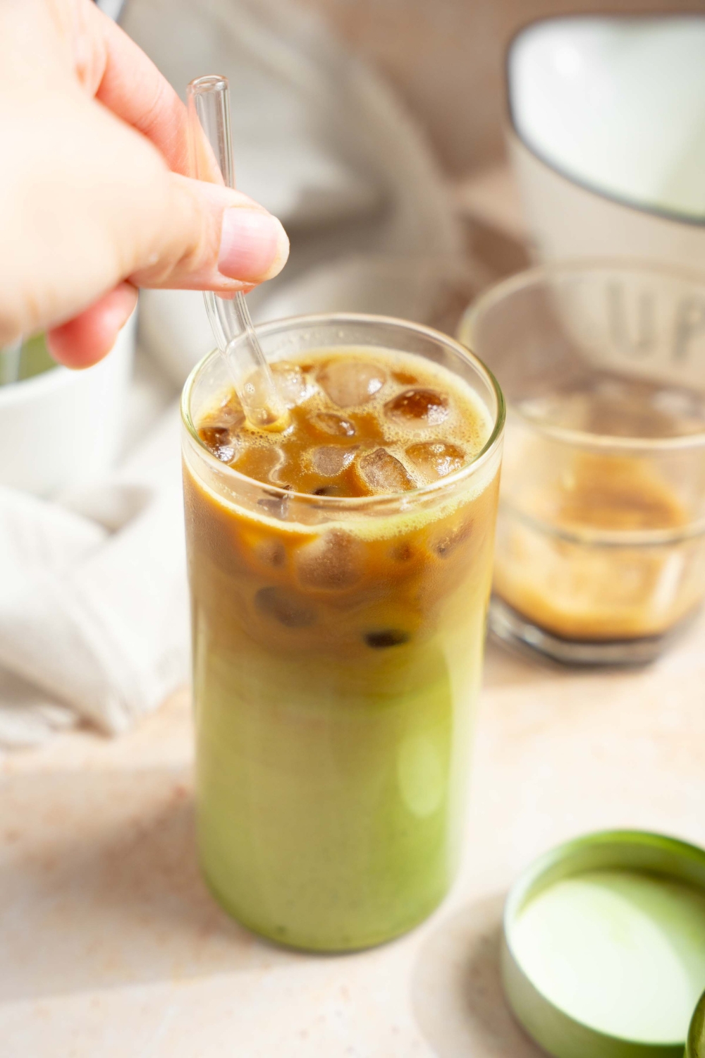 A tall glass of an iced matcha latte with a hand stirring the glass straw in the glass. The glass is on a tan counter with a small poured espresso and tin of matcha powder.