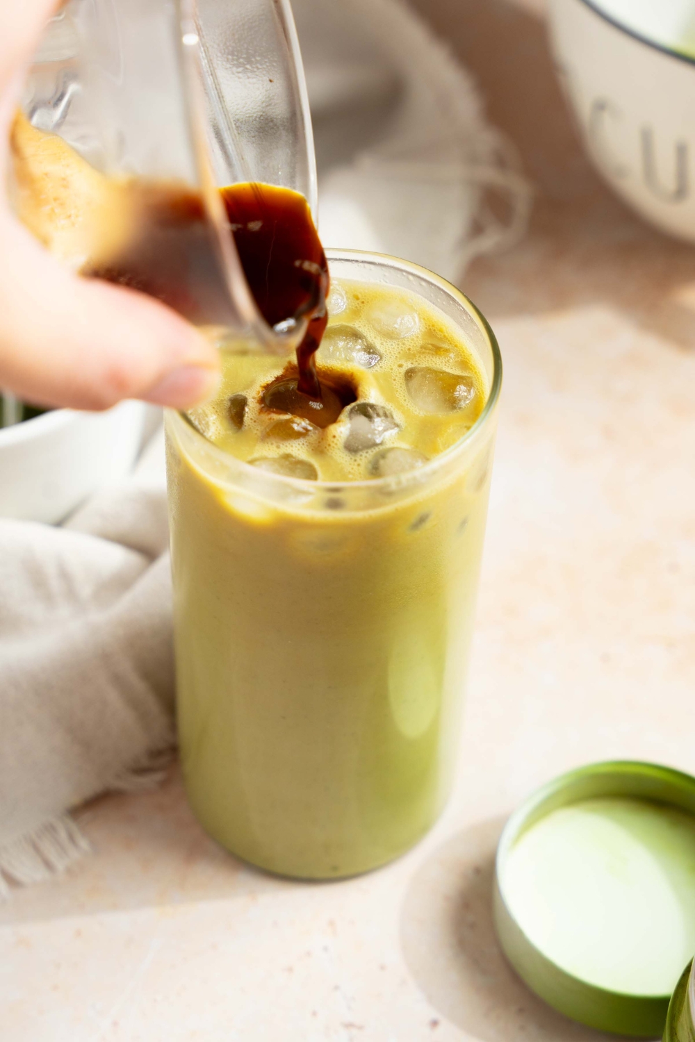 A hand pouring a small glass of espresso into a tall glass of iced matcha latte. The glass is on a tan counter with a white cloth napkin.