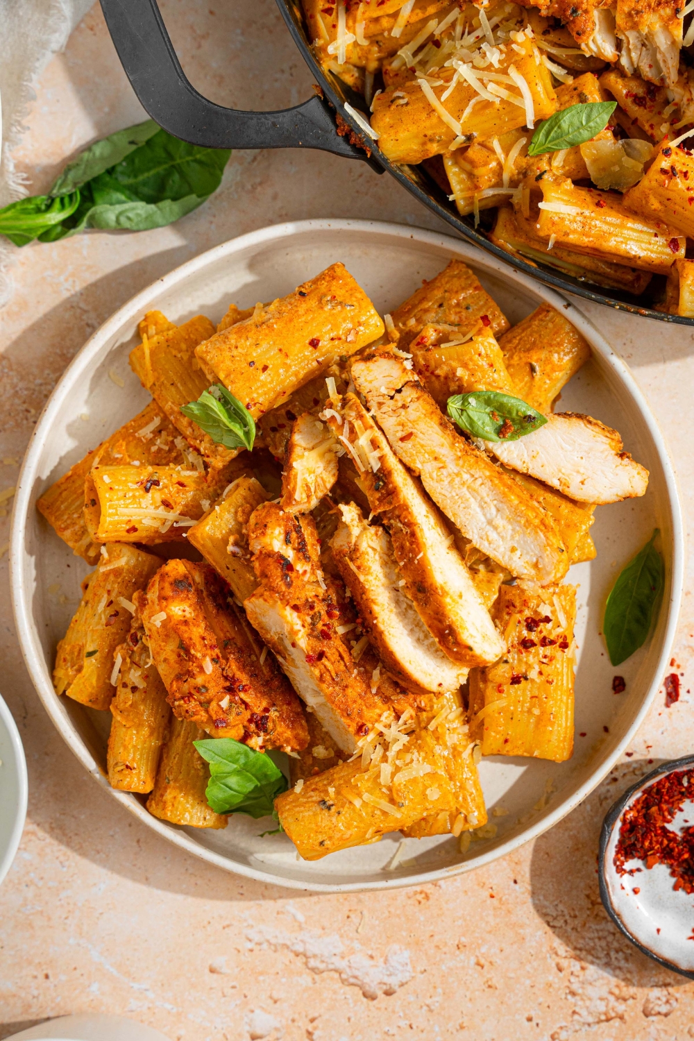 A white plate with garlic parmesan chicken pasta garnished with fresh basil and shredded parmesan. The plate is on a tan counter with a skillet of pasta and small plate of garnishes.