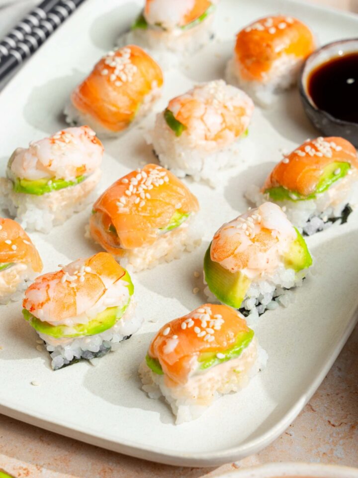 A bunch of pieces of ice cube tray sushi lined up in, rose on a white tray with a cup of soy sauce.